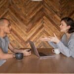 Focused woman explaining opinion to ethnic male coworker during business teamwork sitting at table with laptop and coffee cups in cozy kitchen against wooden wall and looking at each other