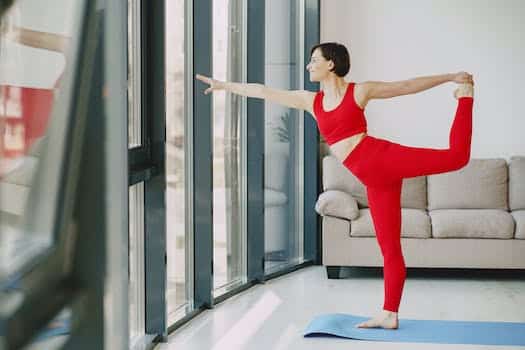 Side view of smiling female in sportswear standing on yoga mat doing Natarajasanaglassasana near window in living room and stretching body