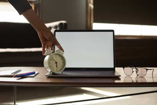 Anonymous black person touching alarm clock near opened netbook with white blank screen placed on table with notebook and pens