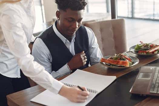 Crop focused diverse coworkers wearing formal outfits taking notes in copybook at table with laptop and dishes during lunch time in modern cafe
