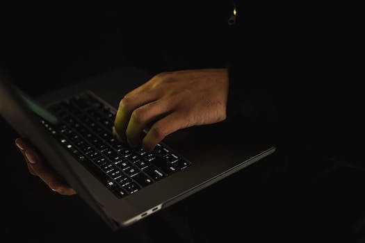 Crop unrecognizable male in black sweater holding netbook on hand and typing on keypad in dark room