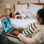 Ethnic mother using laptop near baby sleeping on bed with father