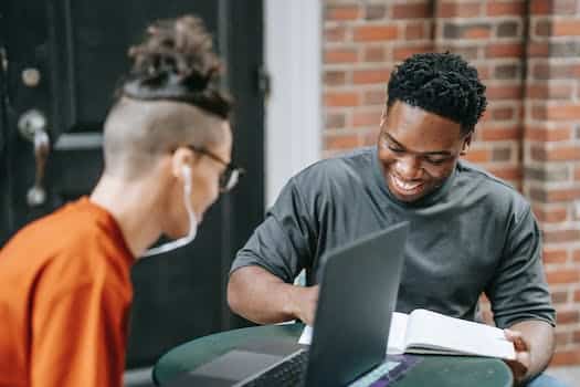 Happy young African American male student smiling and taking notes in copybook while preparing for exams with friend using laptop in street cafe