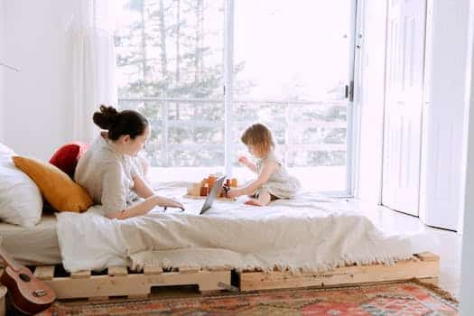 Side view of young focused female in casual clothes lying in bed and using laptop while daughter playing sitting nearby on sunny morning