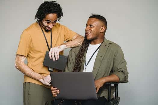 Happy African American androgynous person with Afro braids and computer looking at black colleague with vitiligo and notebook while working on gray background