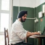 Side view low angle of focused young Sikh male in formal wear and turban sitting at table and working on laptop during remote job