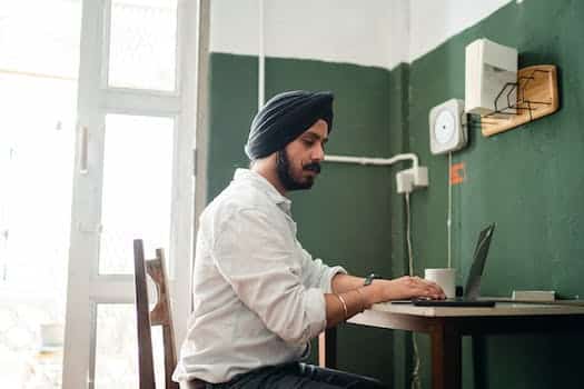 Side view low angle of focused young Sikh male in formal wear and turban sitting at table and working on laptop during remote job