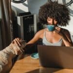 Concentrated young black woman working remotely on netbook while sitting in room at table and drinking coffee while petting dog