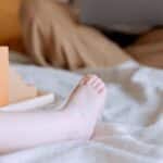 Crop faceless toddler sitting on bed with colorful wooden building blocks while mother sitting with legs crossed doing remote work with laptop on knees on background