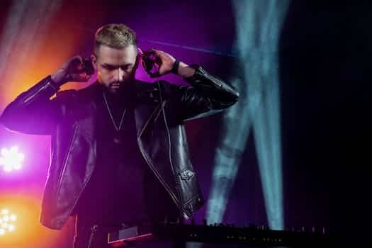 Self assured young bearded male DJ in trendy leather jacket adjusting headphones while performing music on stage in dark illuminated nightclub