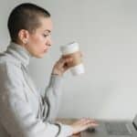 Side view of concentrated female in casual clothes sitting with laptop at table while drinking coffee