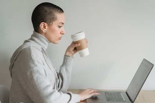 Side view of concentrated female in casual clothes sitting with laptop at table while drinking coffee