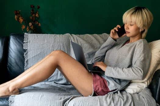 Side view of young ethnic female remote employee speaking on cellphone and surfing internet on netbook while lying on cozy sofa at home