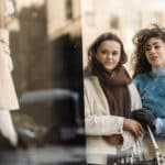 Through glass of stylish young multiracial ladies in trendy warm clothes standing near fashion boutique showcase and choosing clothes