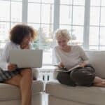 Cheerful young multiethnic girlfriends discussing project while working on netbook at home