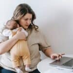 Concentrated young female freelancer embracing newborn while sitting at table and working remotely on laptop at home