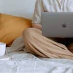 Crop faceless female in casual outfit sitting on bed with legs crossed holding laptop on knees with cup of coffee standing on notebook while working from home