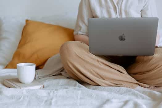 Crop faceless female in casual outfit sitting on bed with legs crossed holding laptop on knees with cup of coffee standing on notebook while working from home
