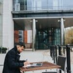 Ethnic distance employee writing in copybook near laptop at table