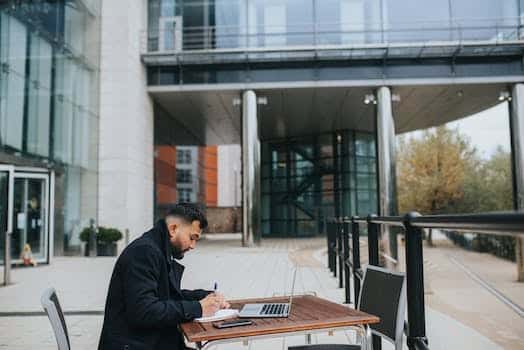 Ethnic distance employee writing in copybook near laptop at table