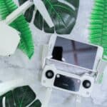 From above of black screen of smartphone placed above flight controller near drone on desk decorated with tropical plants during daylight