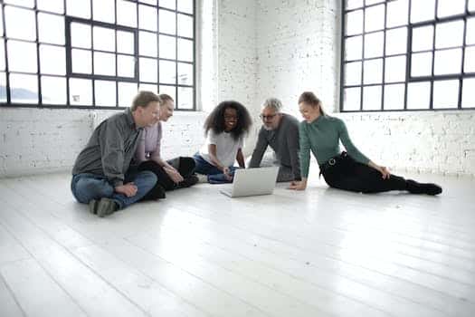 Group of happy multiracial coworkers in casual clothing smiling while watching video on computer gathering on floor in modern workplace