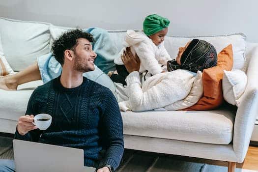 Happy young ethnic man in casual clothes drinking coffee and working on laptop while sitting on floor near Muslim wife lying on sofa and playing with cute toddler daughter
