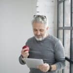 Middle aged bearded man in casual wear with eyeglasses on head watching video on tablet while having break with apple near window