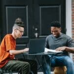 Positive young diverse male colleagues in casual clothes smiling and using laptop while working remotely on project sitting at table in street cafe