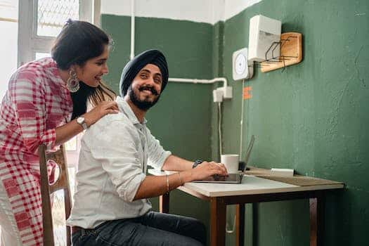 Smiling young Indian woman in casual clothes caring for positive husband working on laptop from home
