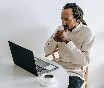 Concentrated adult African American male in formal wear sitting at table with cup of coffee and watching video on netbook in light room