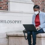 Concentrated young African American female student in casual clothes and medical mask sitting near Philosophy faculty and typing on laptop
