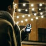 Selective Focus Photography of Man Using Smartphone Beside String Lights