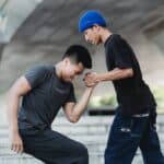 Side view of cool young guy in blue beanie helping best friend in sportswear to stand up while training together in skate park