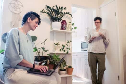 Young woman with short hair standing near wall with cup of coffee and communicating with female friend working remotely on laptop at home